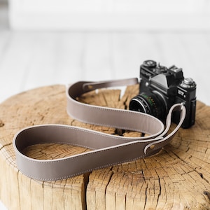 Peut inclure: Une sangle d'appareil photo en cuir taupe avec des coutures blanches et des rivets dor&eacute;s est pos&eacute;e sur une surface en bois. Un appareil photo vintage noir avec un objectif est &eacute;galement sur la surface en bois.