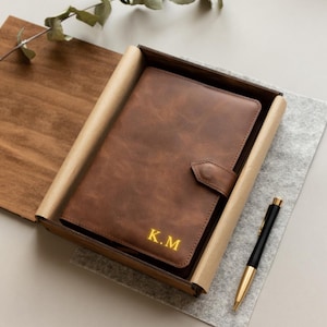 May include: A brown leather journal with the initials "K.M" embossed in gold, presented in a wooden box with a pen. The journal has a clasp closure. The pen is black and gold.