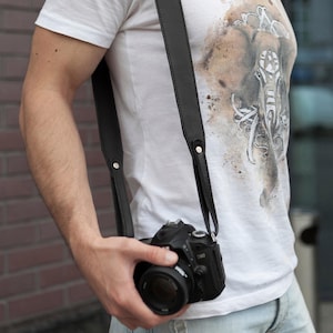Peut inclure: Sangle d'appareil photo en cuir noir avec rivets argent&eacute;s. La sangle est drap&eacute;e sur l'&eacute;paule et tient un appareil photo reflex num&eacute;rique noir avec un objectif. La personne porte un t-shirt blanc avec un motif graphique.
