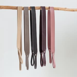 May include: A collection of six leather camera straps in various colors, including beige, taupe, black, burgundy, and mauve. Each strap is suspended from a wooden branch and features a loop and metal hardware for attachment.