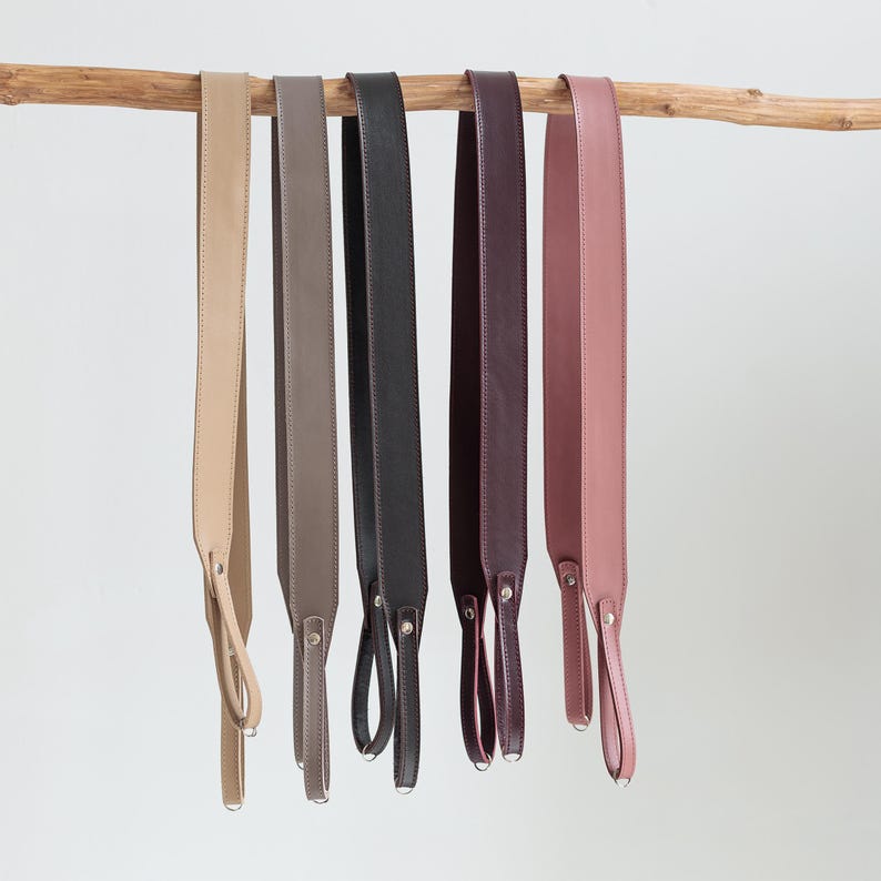 May include: A collection of six leather camera straps in various colors, including beige, taupe, black, burgundy, and mauve. Each strap is suspended from a wooden branch and features a loop and metal hardware for attachment.