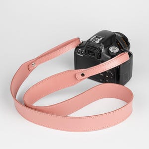 May include: A black DSLR camera with a pink leather camera strap. The strap is a light shade of pink and is attached to the camera with metal hardware. The camera is positioned on a white background.