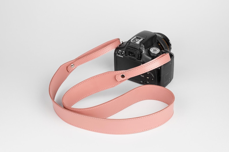 May include: A black DSLR camera with a pink leather camera strap. The strap is a light shade of pink and is attached to the camera with metal hardware. The camera is positioned on a white background.