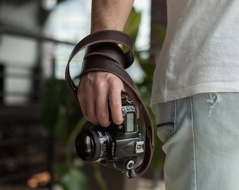 May include: A person holding a black camera with a brown leather strap. The camera has a lens with a silver ring and a digital display.