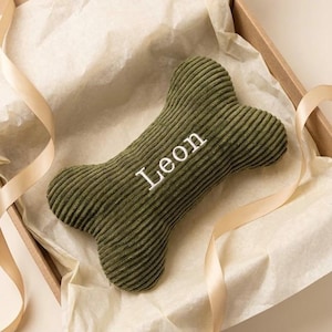 May include: An olive green corduroy dog toy shaped like a bone with the name Leon embroidered in white thread across the centre. The toy is placed inside a cardboard box lined with light beige tissue paper and accented with light beige ribbons.