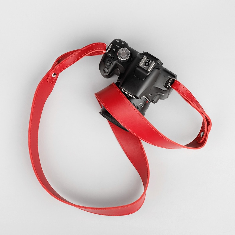 May include: A black Canon camera with a bright red leather camera strap. The strap is looped around the camera body and extends in a curved shape. The camera strap is made of smooth leather and has a simple design.