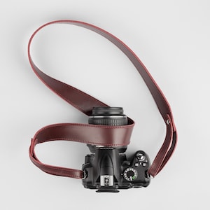 May include: A black DSLR camera with a lens and a burgundy leather camera strap. The strap is wrapped around the camera body and lens. The camera is on a white background.