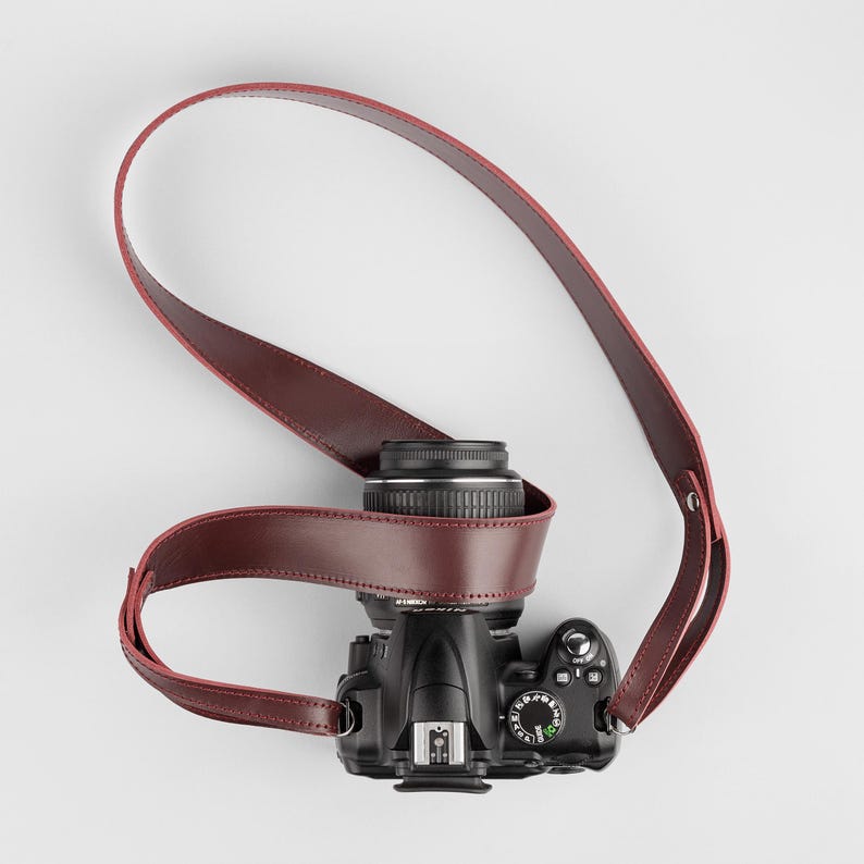 May include: A black DSLR camera with a lens and a burgundy leather camera strap. The strap is wrapped around the camera body and lens. The camera is on a white background.