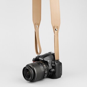 May include: A black Nikon D3000 DSLR camera with a lens attached, suspended by a tan leather camera strap. The strap is attached to the camera with metal rivets. The camera lens is black and has a silver ring.