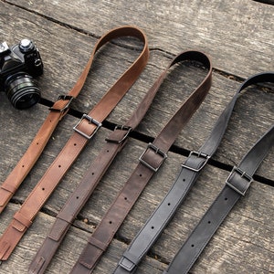 May include: Four leather camera straps in brown and black with metal buckles. The straps are laid out on a wooden surface.