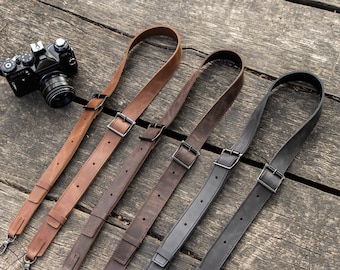 May include: Four leather camera straps in brown and black with metal buckles. The straps are laid out on a wooden surface.