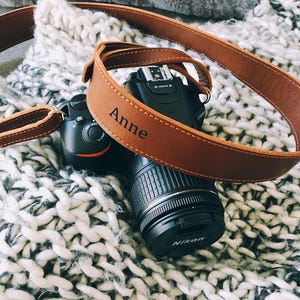 Peut inclure: Un appareil photo reflex num&eacute;rique Nikon noir avec une sangle en cuir marron. La sangle est personnalis&eacute;e avec le nom "Anne" et est drap&eacute;e sur l'appareil photo. L'appareil photo et la sangle reposent sur une couverture en tricot blanc et gris.