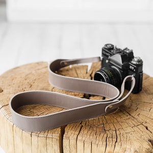 May include: A taupe leather camera strap with a black camera on a wooden surface. The strap is looped and has a simple, elegant design. The camera is a vintage model.