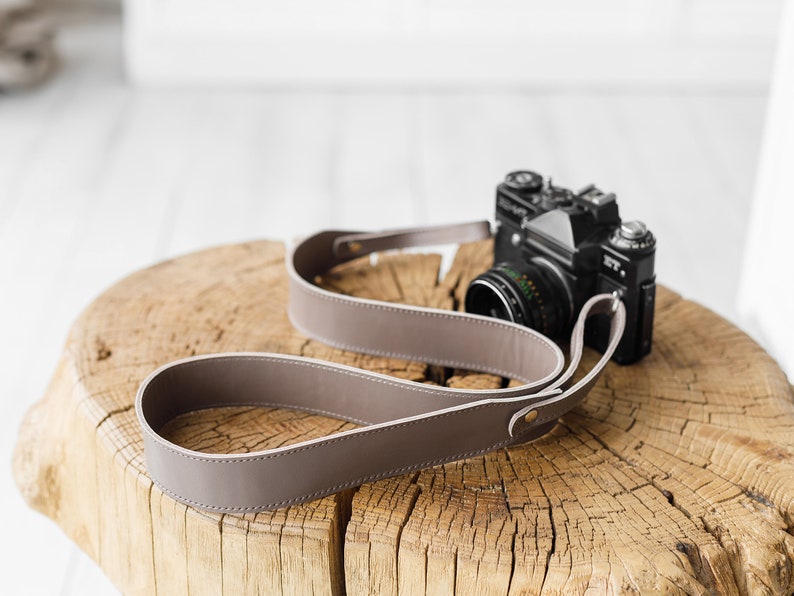May include: A taupe leather camera strap with a black camera on a wooden surface. The strap is looped and has a simple, elegant design. The camera is a vintage model.