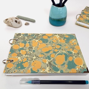May include: A ring-bound notebook with a marbled design in teal, orange, and grey. The notebook is open, showing the patterned cover. A pen and a small teal vase with pencils are also visible.