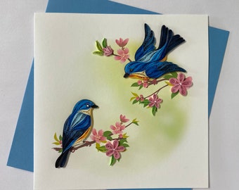 May include: A handmade greeting card featuring two blue birds perched on a branch with pink flowers. The birds are made of paper and have intricate details. The card is on a blue background.