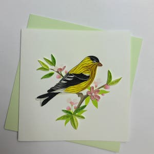 May include: A yellow and black bird with a black head perched on a branch with pink flowers and green leaves. The bird is made of paper and is a paper quilling art piece.