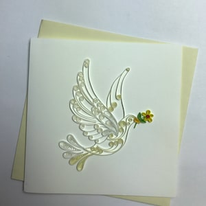 Puede incluir: Tarjeta de felicitación hecha a mano con una paloma blanca en vuelo, elaborada con técnicas de quilling. La paloma sostiene una pequeña flor amarilla y roja en su pico. La tarjeta está sobre un fondo blanco con un sobre crema.