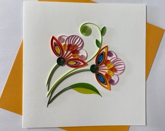 May include: A handmade greeting card featuring two colorful paper flowers with pink, orange, yellow, red, and green accents. The flowers are arranged on a white card with a light orange envelope.