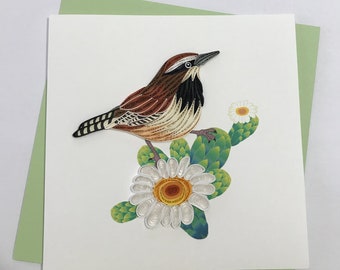 May include: A handmade paper quilling card featuring a brown and white bird perched on a green cactus with white flowers. The card is on a green background.