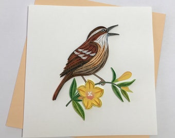 May include: A brown and white paper quilled bird with a yellow flower and green leaves. The bird is singing with its mouth open.