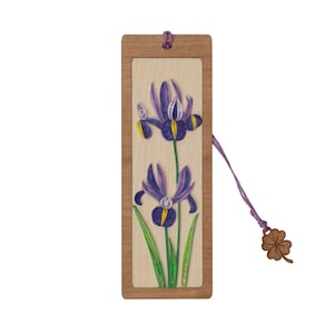 Pode incluir: Um marcador de página de madeira com um design de flor de papel roxo e verde. O marcador tem uma moldura de madeira marrom e um cordão com um amuleto de trevo de quatro folhas.