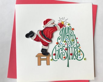 May include: A handmade Christmas card featuring a quilled Santa Claus decorating a Christmas tree. Santa is in a red suit with white trim, standing on a small stool. The tree is green with colorful ornaments, and the words "Merry Christmas" are written in green.
