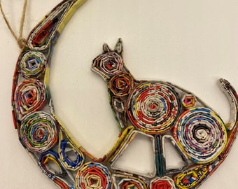May include: A handcrafted cat silhouette ornament perched on a crescent moon, made from rolled paper. The ornament features a variety of colors, including red, yellow, and blue. A twine loop is attached for hanging. The cat and moon are made from recycled materials.