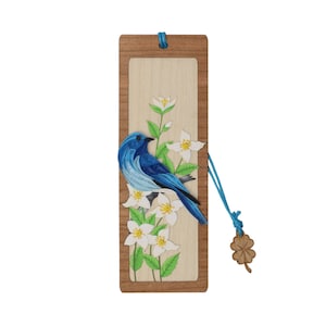 May include: A wooden bookmark with a blue bird and white flowers on a light wood background. The bookmark has a blue ribbon attached to it with a four-leaf clover charm.