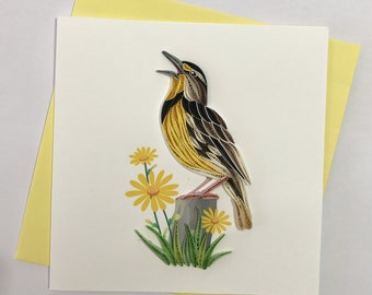 May include: A yellow and brown bird with a long beak is singing on a small wooden stump. The bird is surrounded by yellow flowers with green leaves. The image is a handmade paper quilling art card.