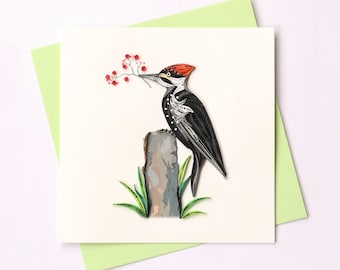May include: A paper quilling card featuring a black and white woodpecker with a red head perched on a tree stump. The woodpecker is looking to the left and has its beak open. There are red berries on a branch behind the woodpecker.