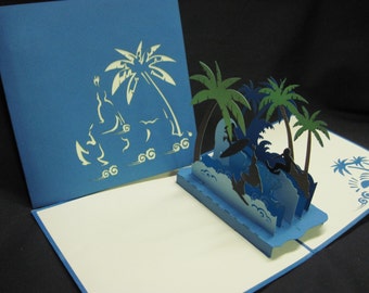 May include: A blue and white pop-up card with a 3D scene of palm trees and surfers riding waves. The card is open and the scene is visible.