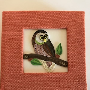 May include: A framed paper quilling artwork of a brown owl perched on a branch with green leaves. The frame is made of a textured orange fabric.