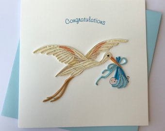 May include: A handmade greeting card with a stork carrying a baby in a blue bundle. The stork is made of paper quilling with tan and orange accents. The card reads "Congratulations" in blue script.