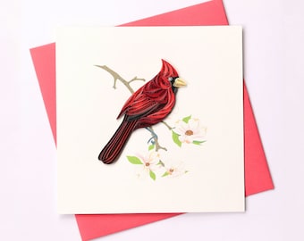 May include: A red cardinal bird with black eyes and a yellow beak perched on a branch with white flowers. The bird is made of paper and is a 3D paper craft.