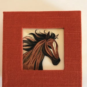 Può includere: Un'opera d'arte in quilling di carta incorniciata che raffigura la testa di un cavallo marrone con una striscia bianca sul viso. La criniera e la coda del cavallo sono fatte di strisce di carta arricciate. La cornice è realizzata in tessuto rosso con una superficie testurizzata.