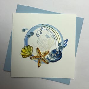 May include: A handmade greeting card with a blue, white, and gold paper quilling design of a seashell, starfish, and a swirl. The card is on a light blue background.