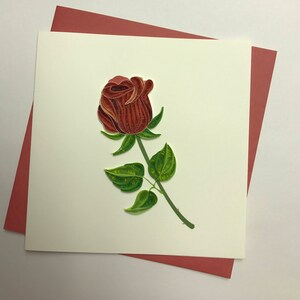 May include: A red paper rose with green paper leaves on a white card with a red border.