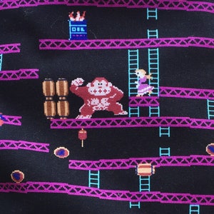 May include: A black zippered pouch with a teal bottom. The pouch has a repeating pattern of a video game level with a monkey, barrels, and ladders. The pouch is sitting on a wooden surface next to a ball of pink and white yarn.