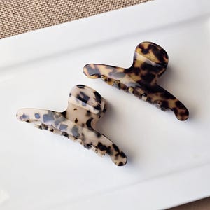 May include: Two tortoiseshell hair claw clips. One clip is a light beige with dark spots, and the other is a darker brown with black spots. The clips are made of a glossy material and are on a white surface.