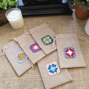 May include: Four rectangular beige burlap drawstring pouches, each featuring a small, square, multi-colored crochet granny square patch centered on the front. The crochet patches display various color combinations including blue, yellow, pink, red, and green.