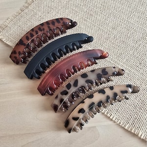 May include: Five banana hair clips in various colors and patterns. The clips include a dark brown leopard print, solid black, reddish-brown, light brown with black spots, and a light tan leopard print. They are arranged on a light brown surface.