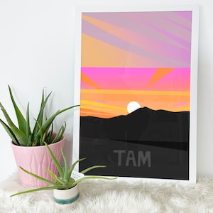 May include: A framed print with a colorful sunset scene over a mountain range. The sky is a gradient of pink, orange, and yellow. The mountain is black and the text "TAM" is printed in white at the bottom of the print.
