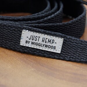 May include: A black hemp dog leash with a woven texture. A small, rectangular tag is attached, displaying the text "JUST HEMP BY WIGGLYWOOS" in a simple, sans-serif font. The leash is coiled on a wooden surface.