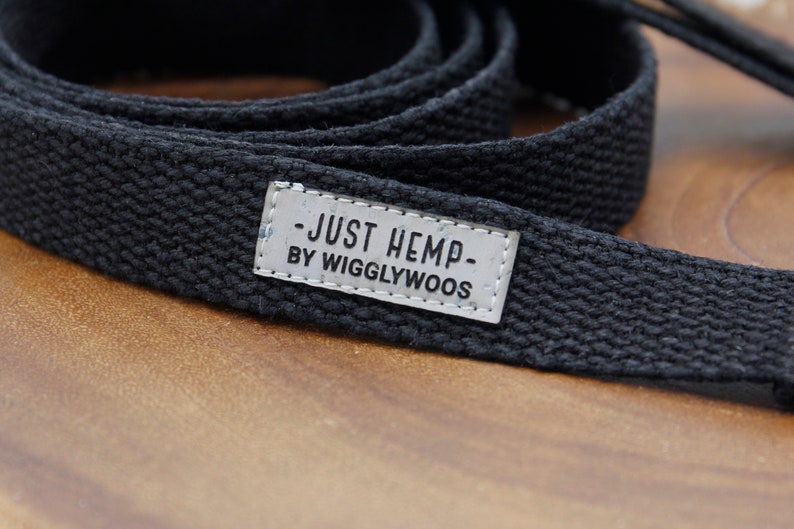 May include: A black hemp dog leash with a woven texture. A small, rectangular tag is attached, displaying the text "JUST HEMP BY WIGGLYWOOS" in a simple, sans-serif font. The leash is coiled on a wooden surface.