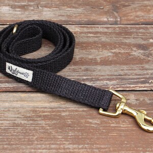 May include: A black dog leash with a gold-colored clasp and a small white label that reads "Wigglywoos" with a paw print. The leash is made of a woven material and is coiled on a wooden surface.