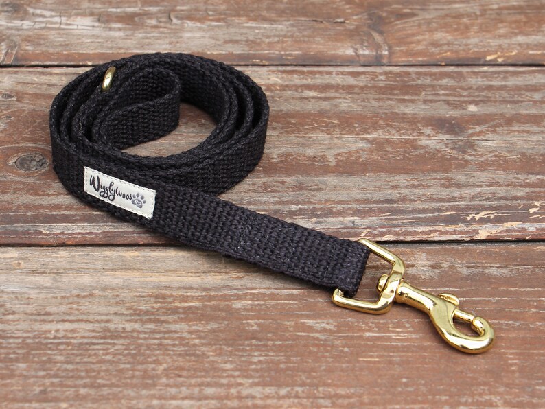 May include: A black dog leash with a gold-colored clasp and a small white label that reads "Wigglywoos" with a paw print. The leash is made of a woven material and is coiled on a wooden surface.