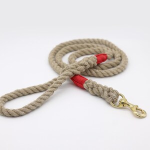 May include: A beige rope dog leash with red accents and a gold-colored clasp. The leash is coiled and lies on a white surface. The rope is thick and appears durable, suitable for pet use.