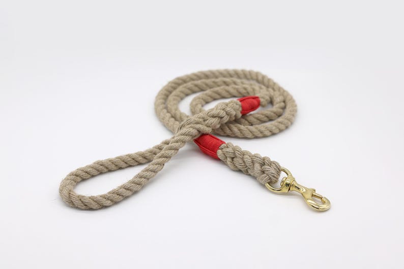 May include: A beige rope dog leash with red accents and a gold-colored clasp. The leash is coiled and lies on a white surface. The rope is thick and appears durable, suitable for pet use.