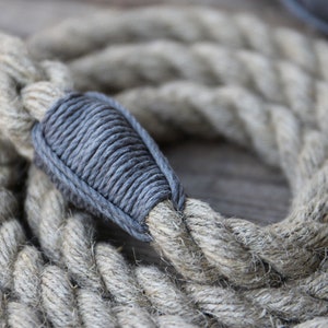 May include: Close-up of a coiled, thick rope with a gray, wrapped section. The rope is a natural beige color, with a textured, braided appearance. The gray section is tightly wound, creating a contrasting detail.